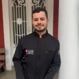 Man in a black chef jacket standing indoors in front of an ornate white door, smiling at the camera.